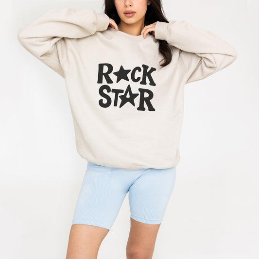 A woman wearing a beige sweatshirt with the text "ROCK STAR" printed on it, standing with her hands behind her head.